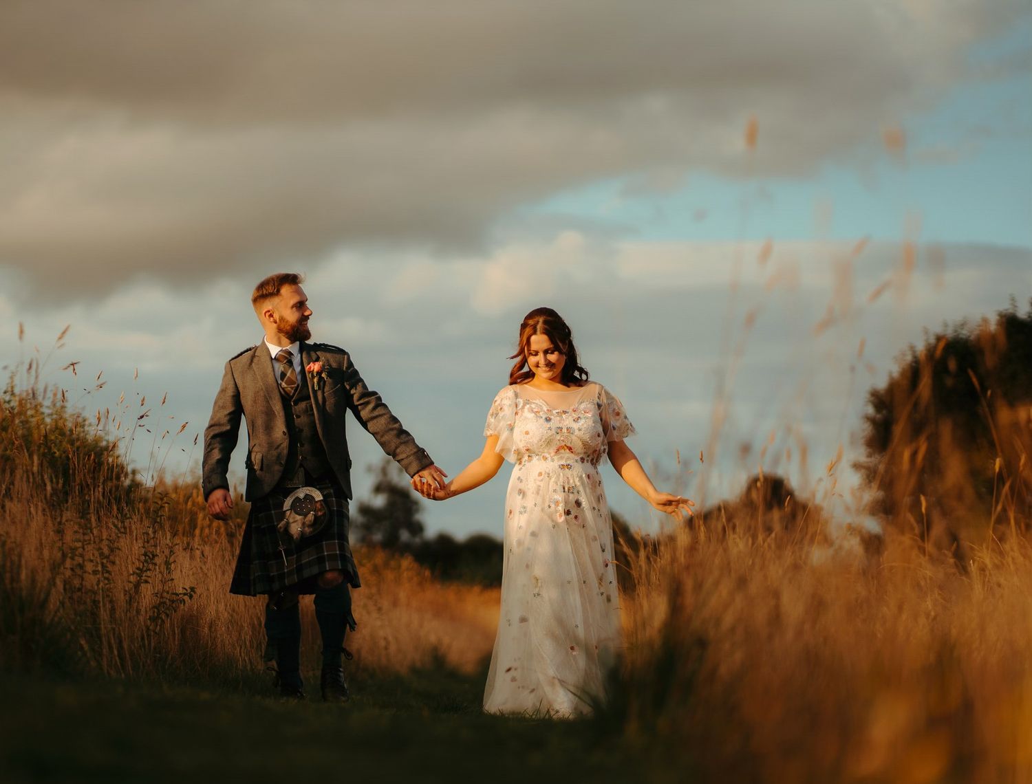 Bride and groom hold hands walking through golden fields in Scotland, groom wearing traditional kilt under dramatic sky.