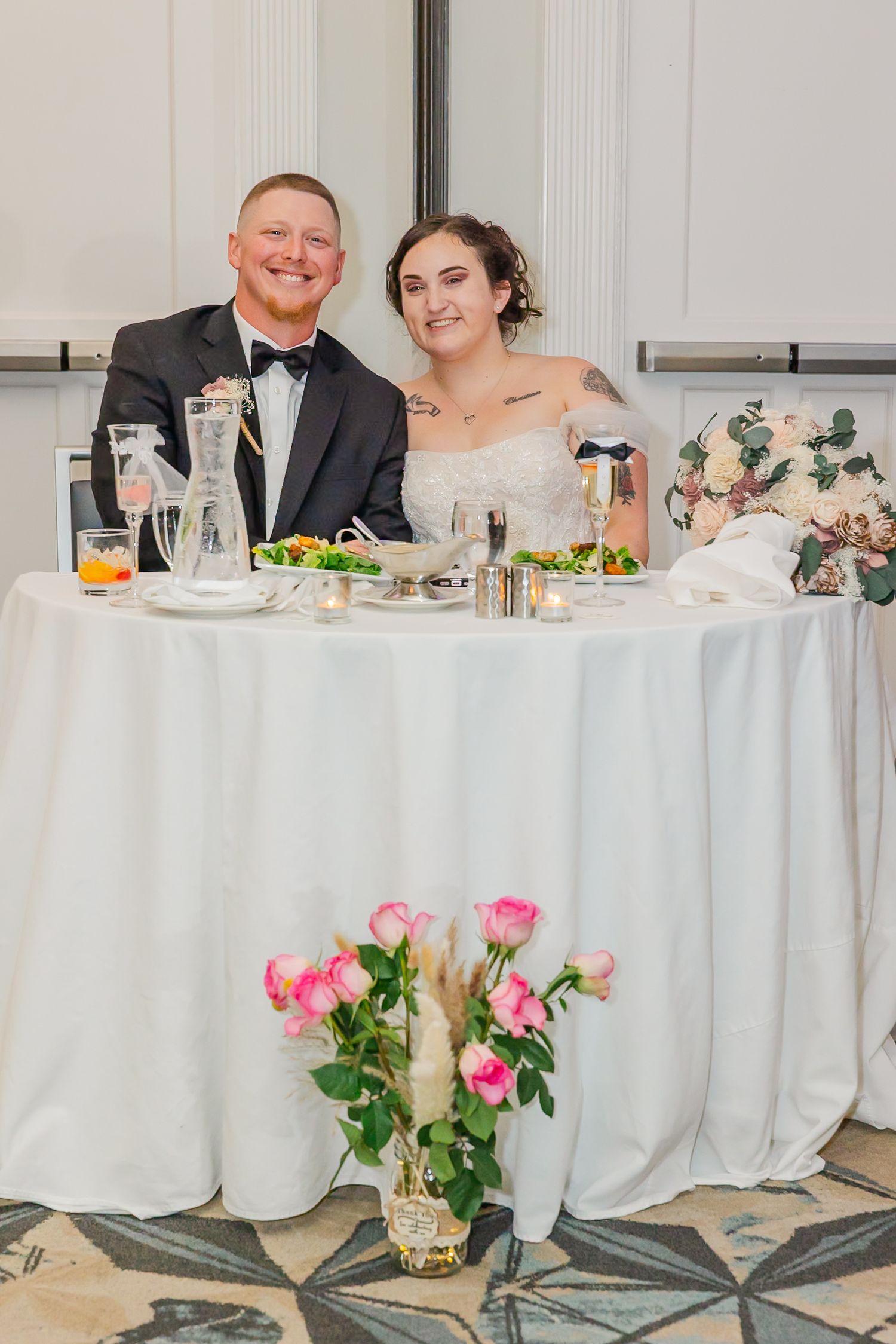 A couple sits at an elegant white-clothed wedding reception table with pink floral centerpieces and table settings.