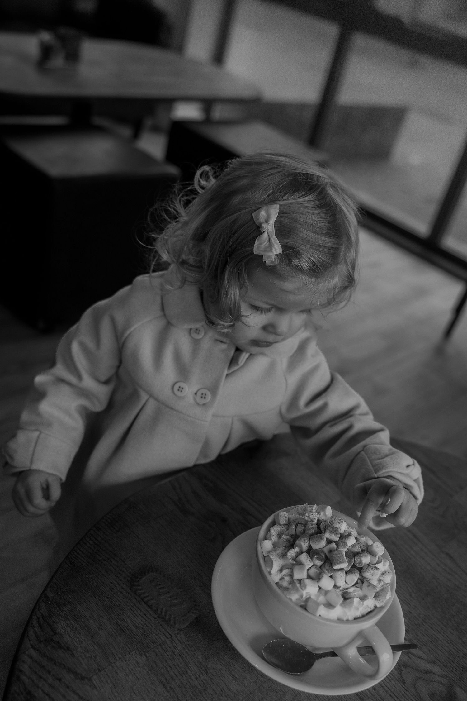 A young child in a light coat sits at a table with a large mug filled with marshmallows in black and white.