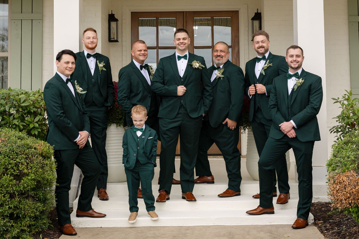 Groomsmen in black tuxedos showing off colorful patterned socks while posing in a row on white porch steps.