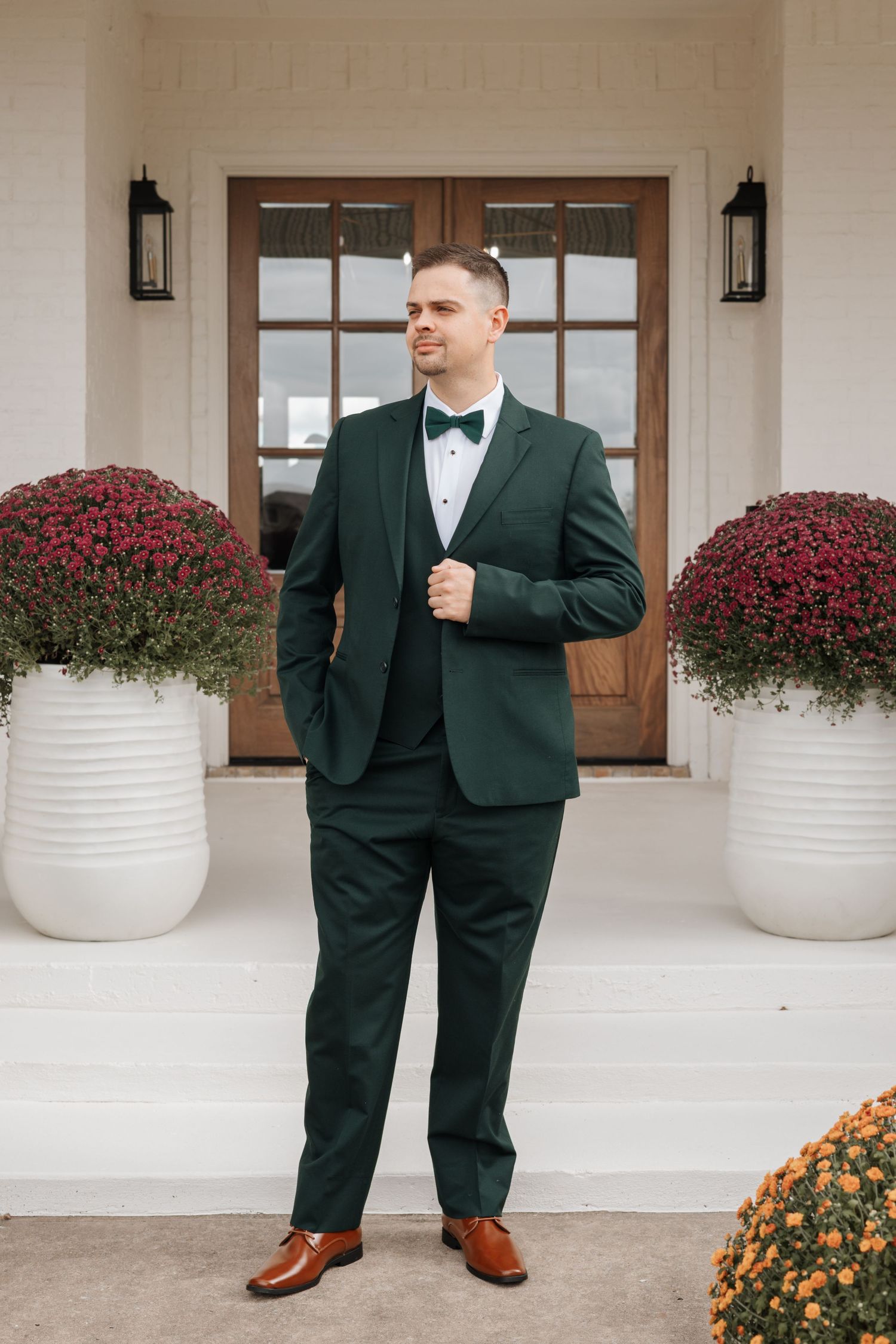 A person in an elegant dark green suit and bow tie stands between two white planters with burgundy flowers.