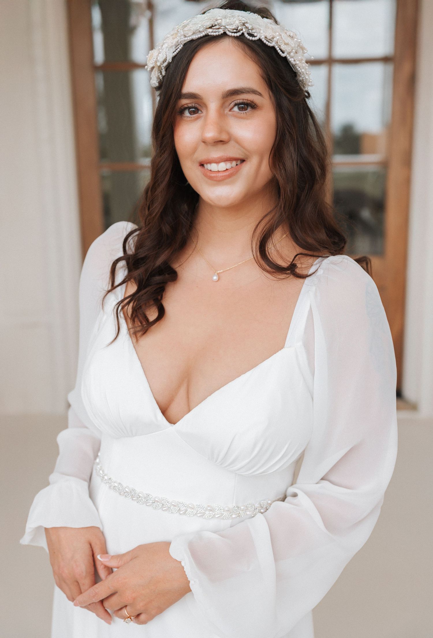 A bride in a white wedding dress with flowing sleeves poses elegantly in front of a window.
