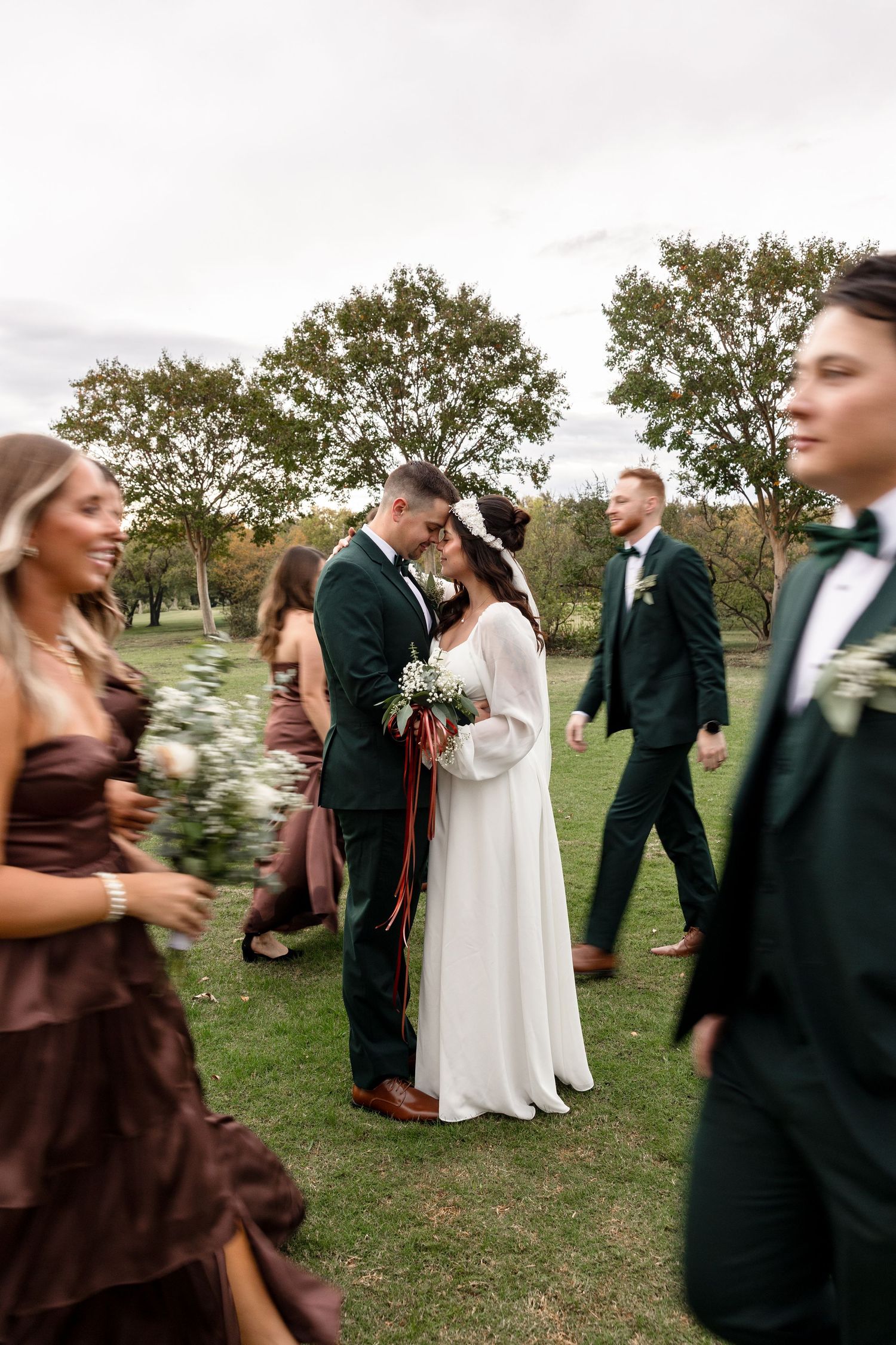 Wedding party members in brown and black attire dance and celebrate on a grassy lawn during an outdoor reception.