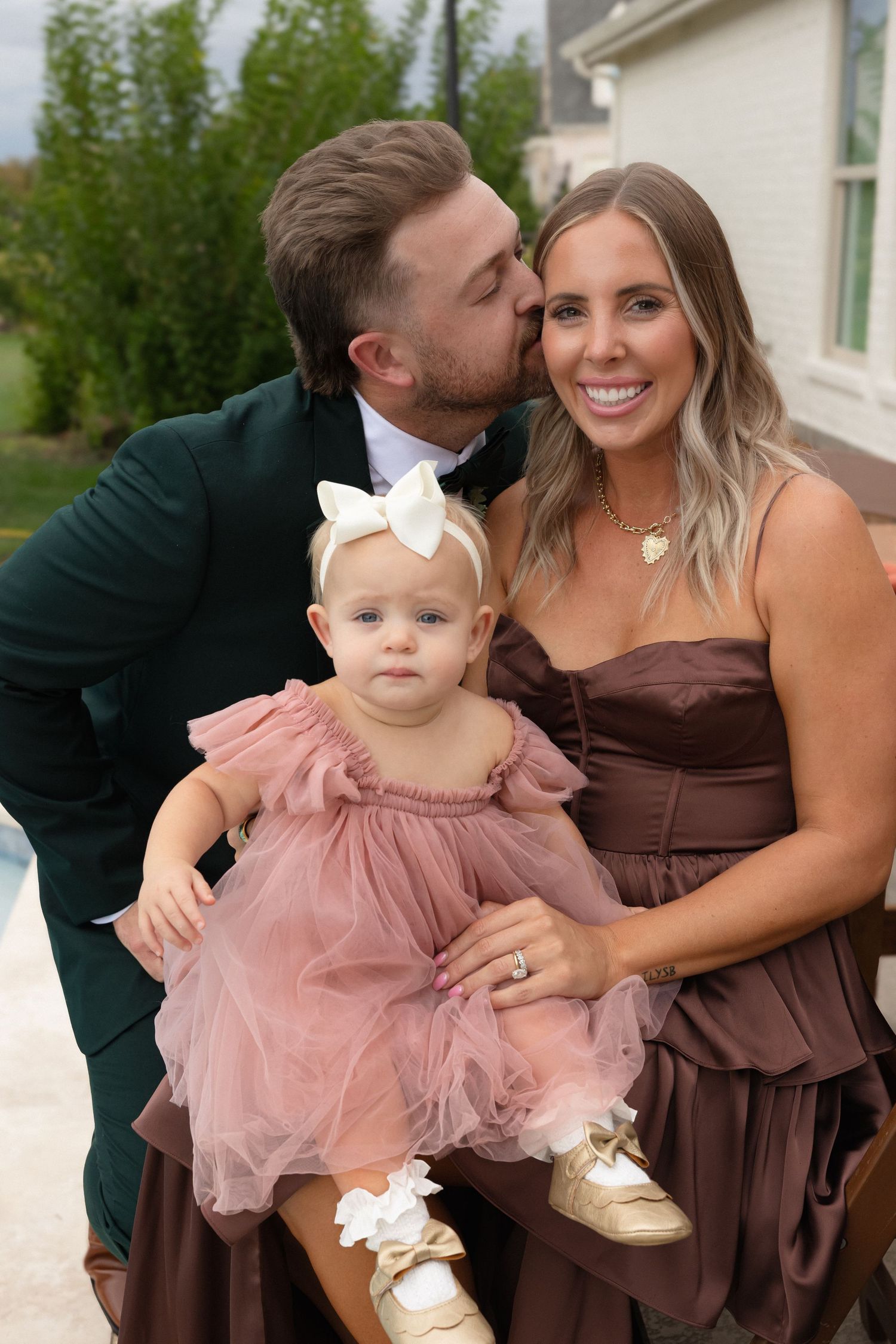 Family dressed in formal attire poses together outdoors, with baby wearing pink tulle dress and white bow headband.