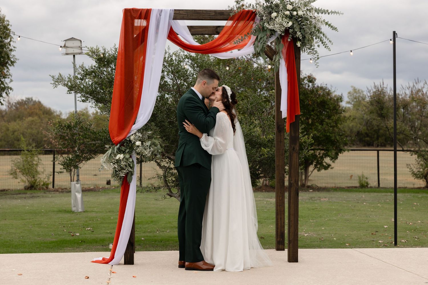 A couple shares a romantic kiss under a wooden arch decorated with red and white draping fabric at an outdoor wedding ceremony.