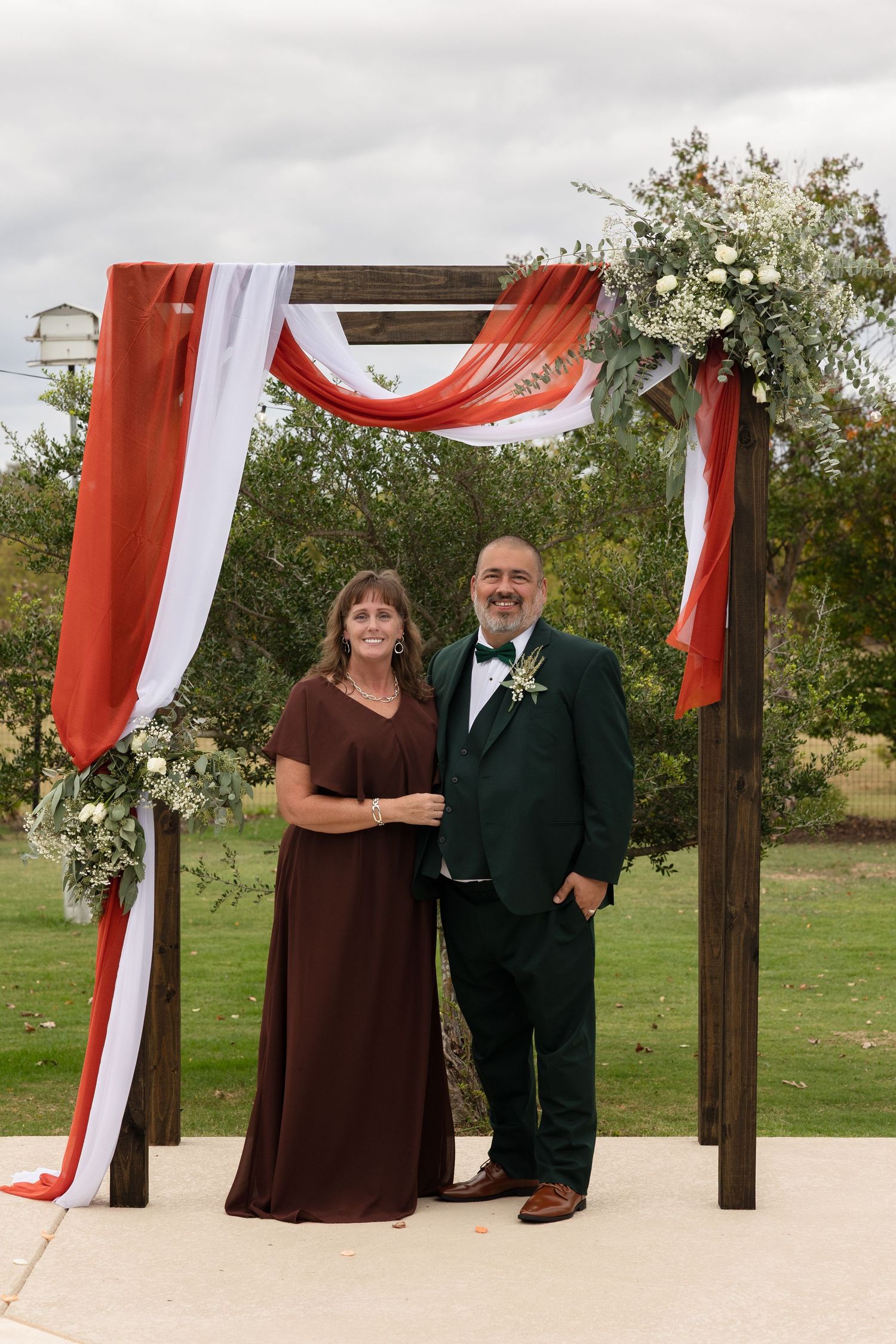 A couple stands under a wooden arch decorated with red and white draping fabric and floral arrangements at an outdoor venue.
