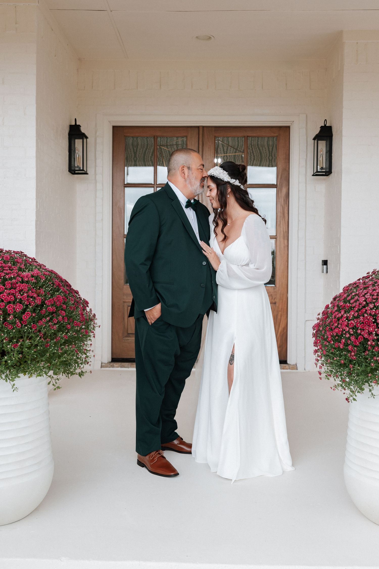 A couple in wedding attire shares an intimate moment on a white porch with pink flowers in decorative planters.
