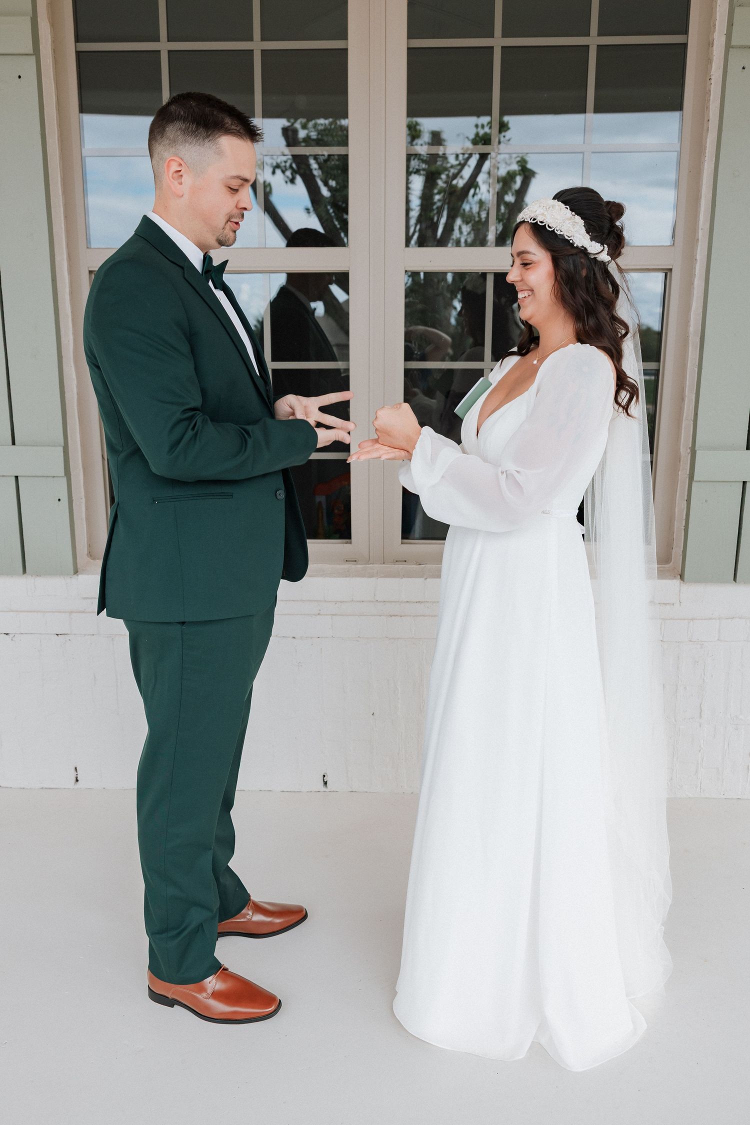 Bride and groom share a playful first look moment on their wedding day in front of white shuttered windows.