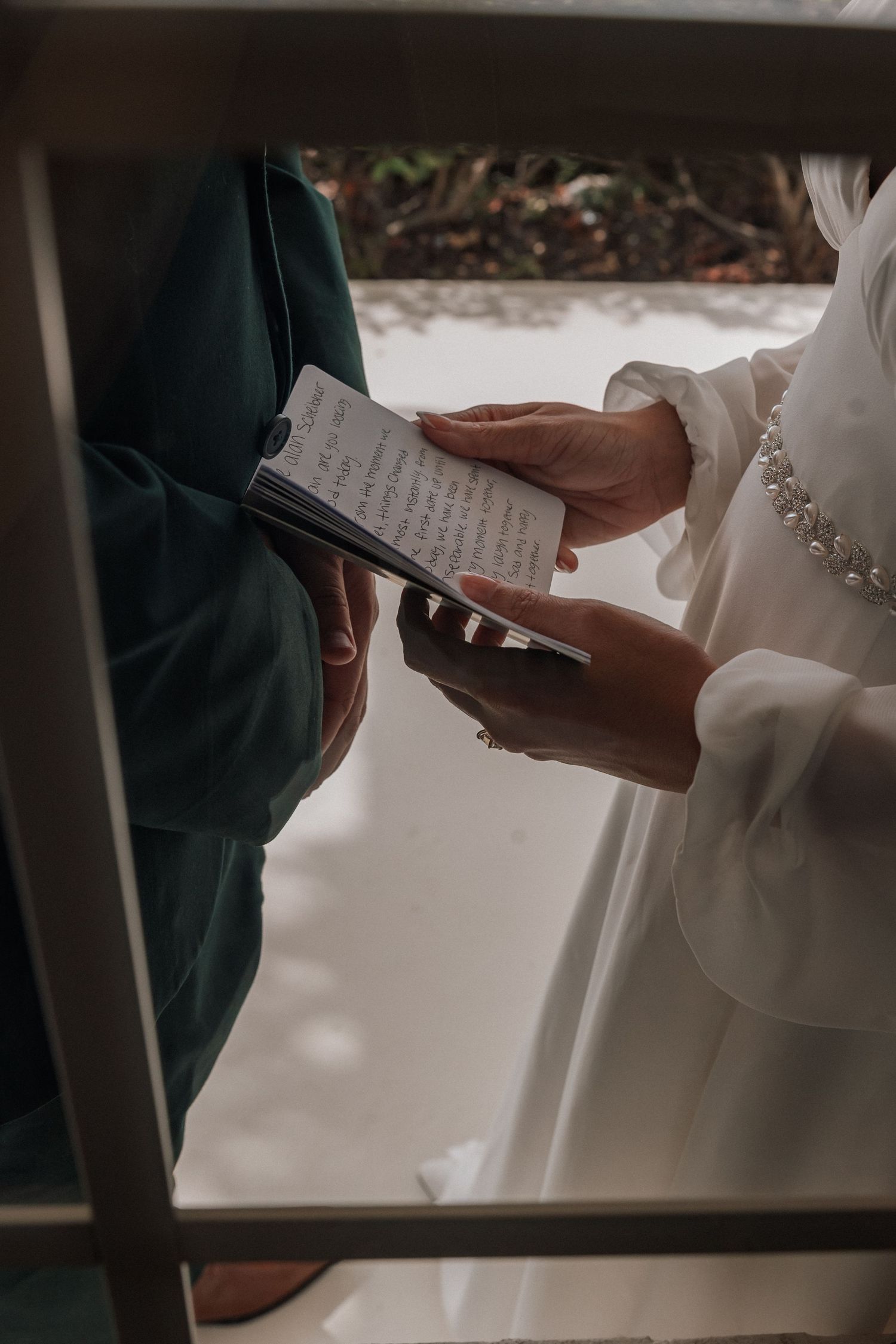 Hands hold handwritten wedding vows during an intimate ceremony viewed through a window frame.