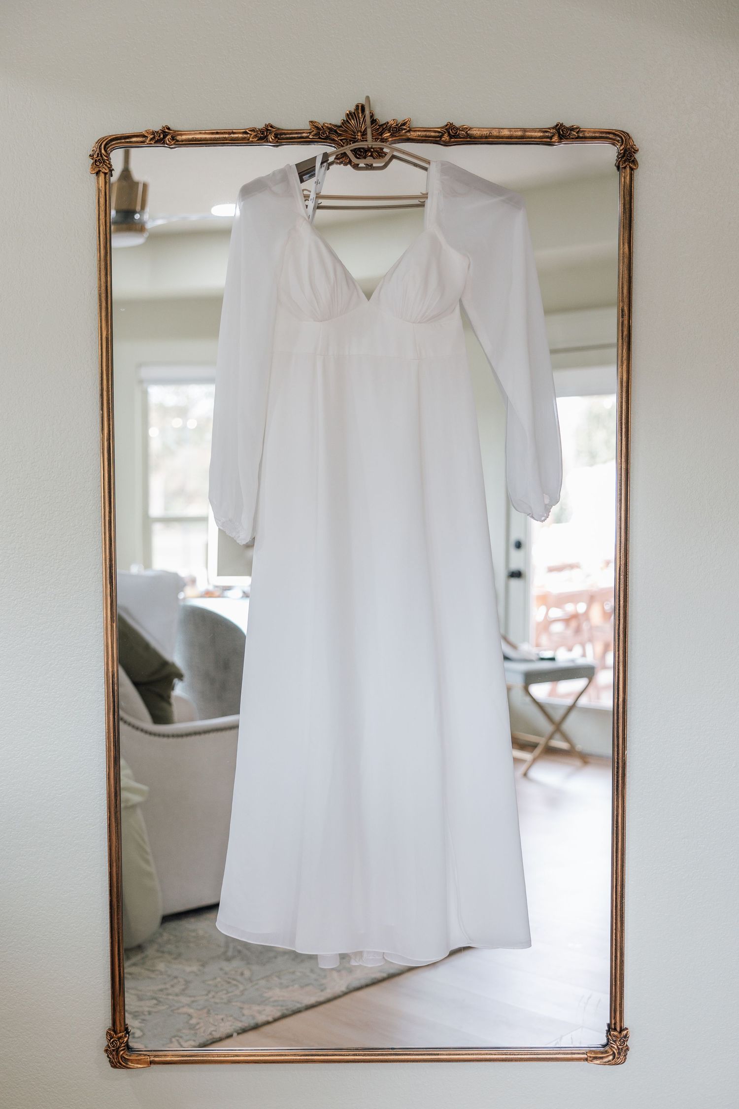 A white flowing dress with long sleeves hangs on a copper-framed mirror in a bright interior space.