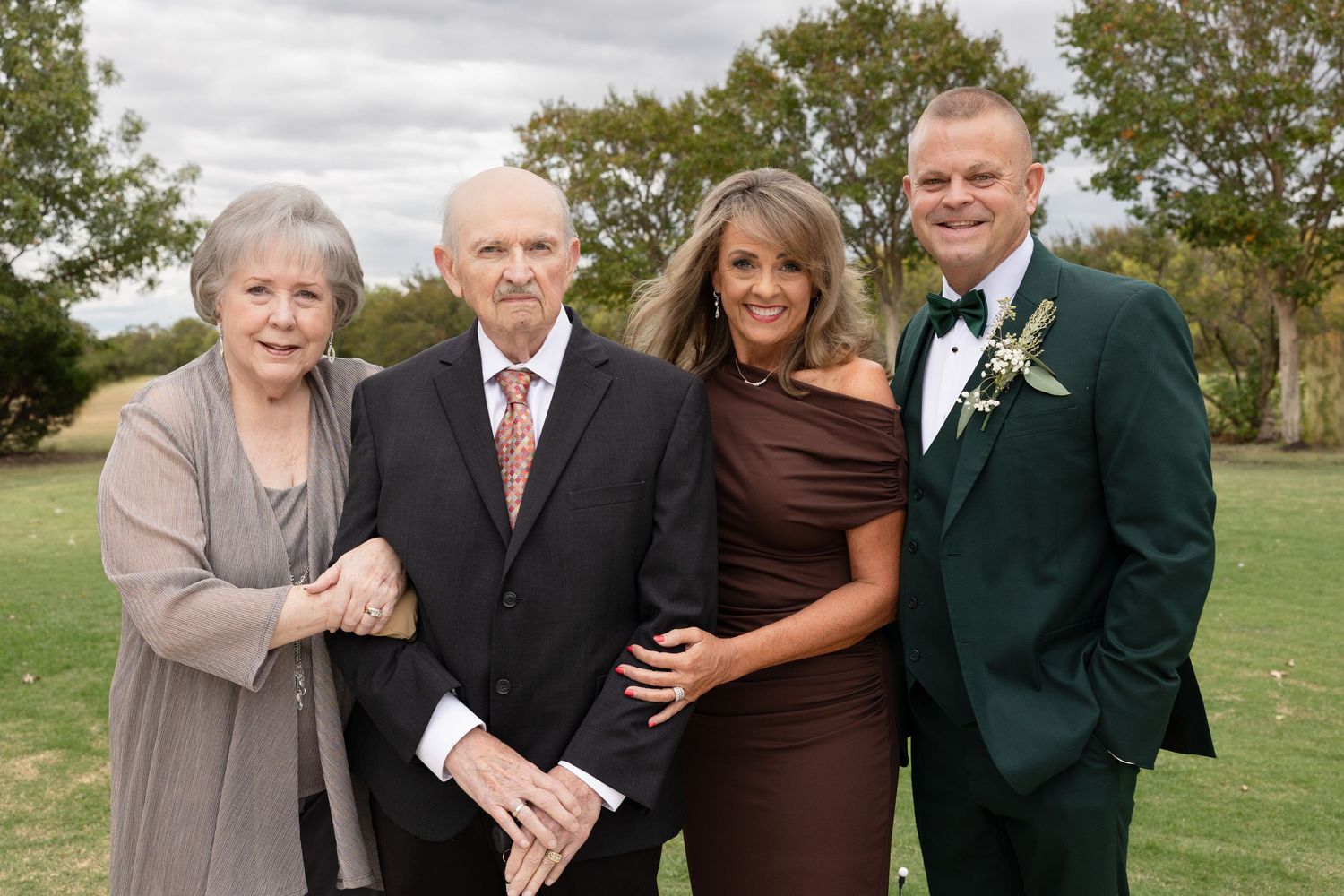 Four people in formal attire pose together outdoors on a grassy area with trees in the background.