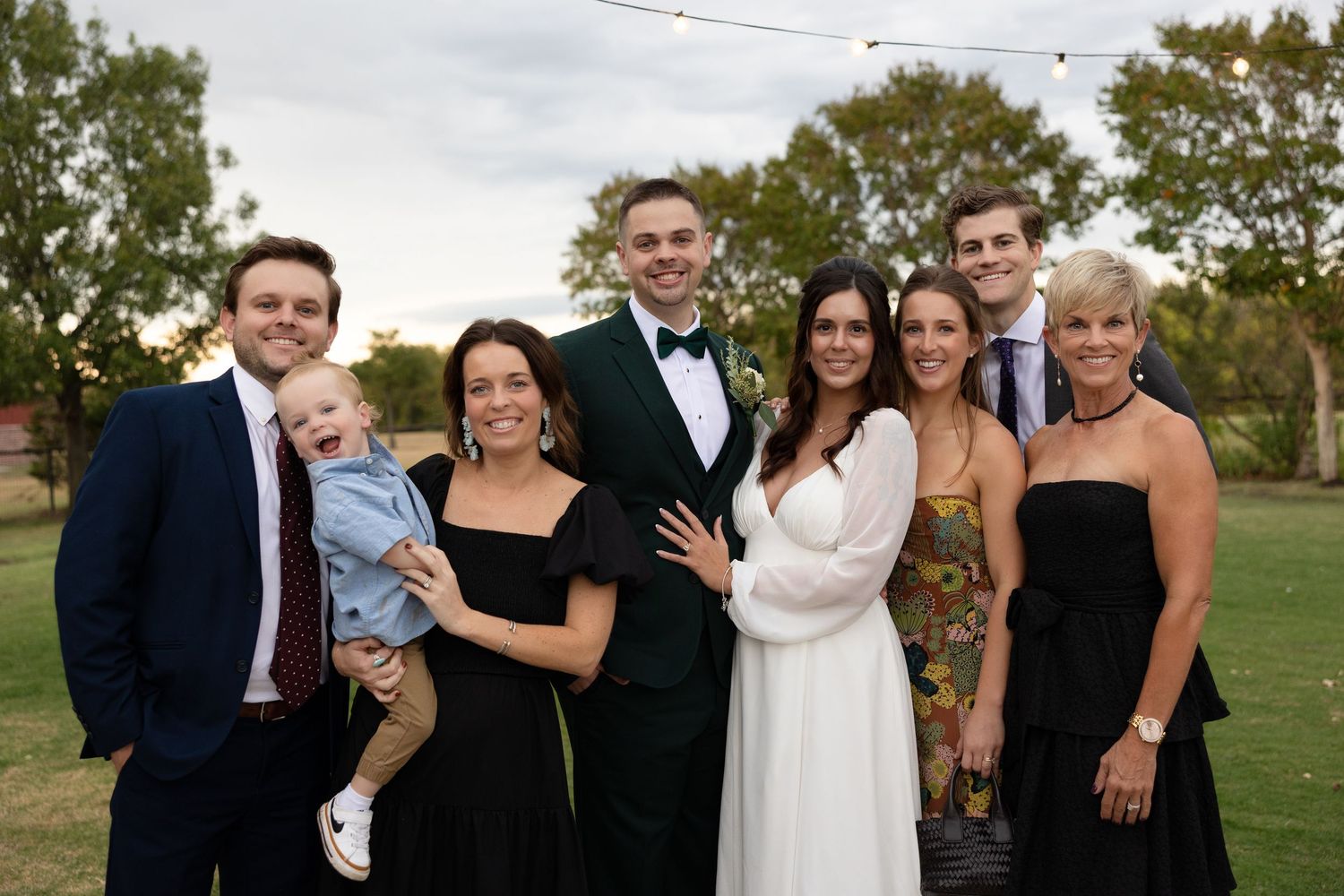 A group of wedding party members pose together outdoors on a grassy lawn during a celebration.