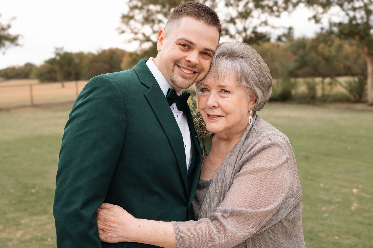 A couple in formal attire share a joyful embrace while posing outdoors in a grassy field.