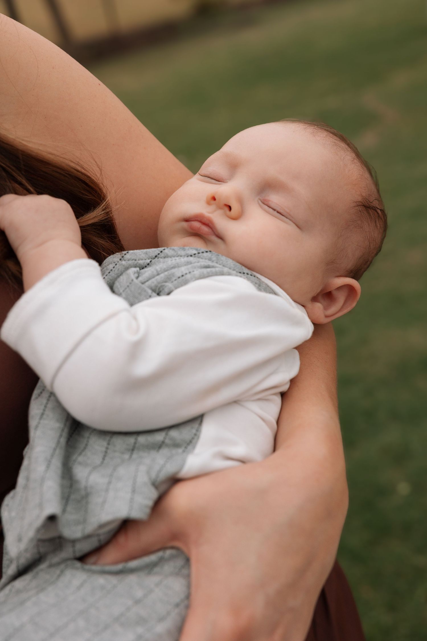 A sleeping baby in a white onesie rests peacefully against someone's shoulder in an outdoor setting.