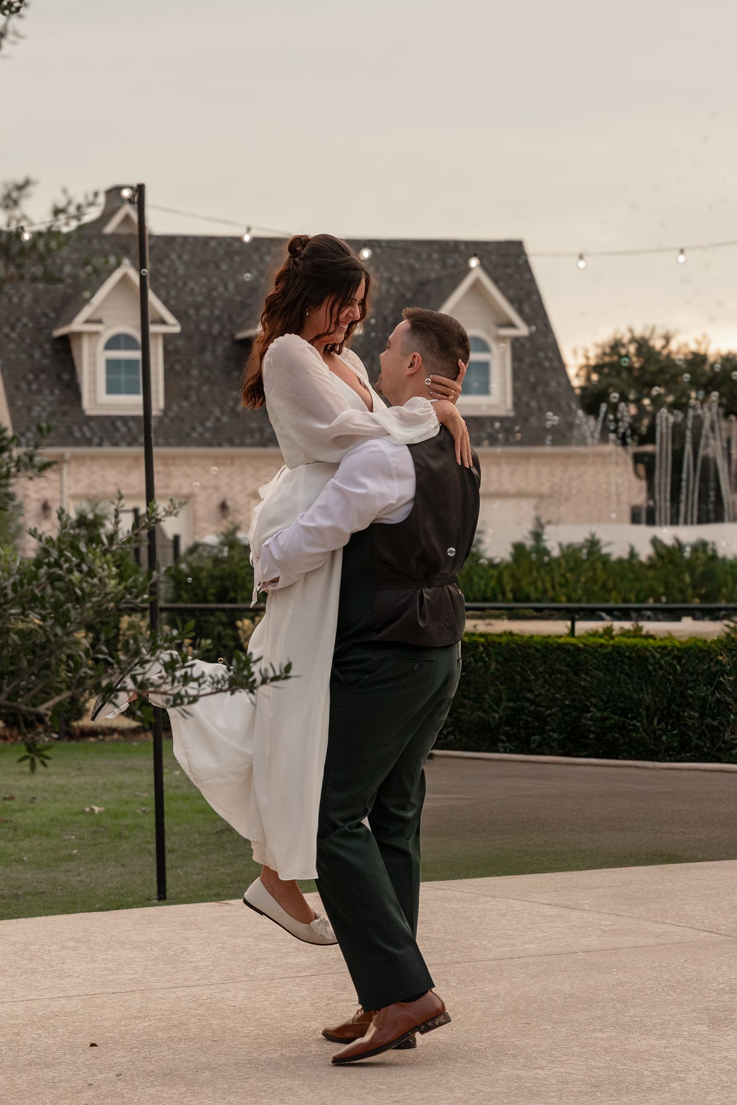A couple in formal attire dancing outdoors with houses in the background during sunset.
