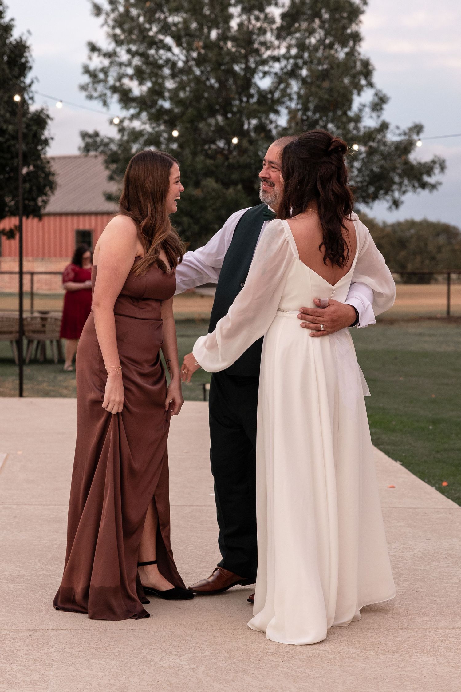 Three people share an intimate moment at an outdoor wedding ceremony on a concrete path near a rustic building.