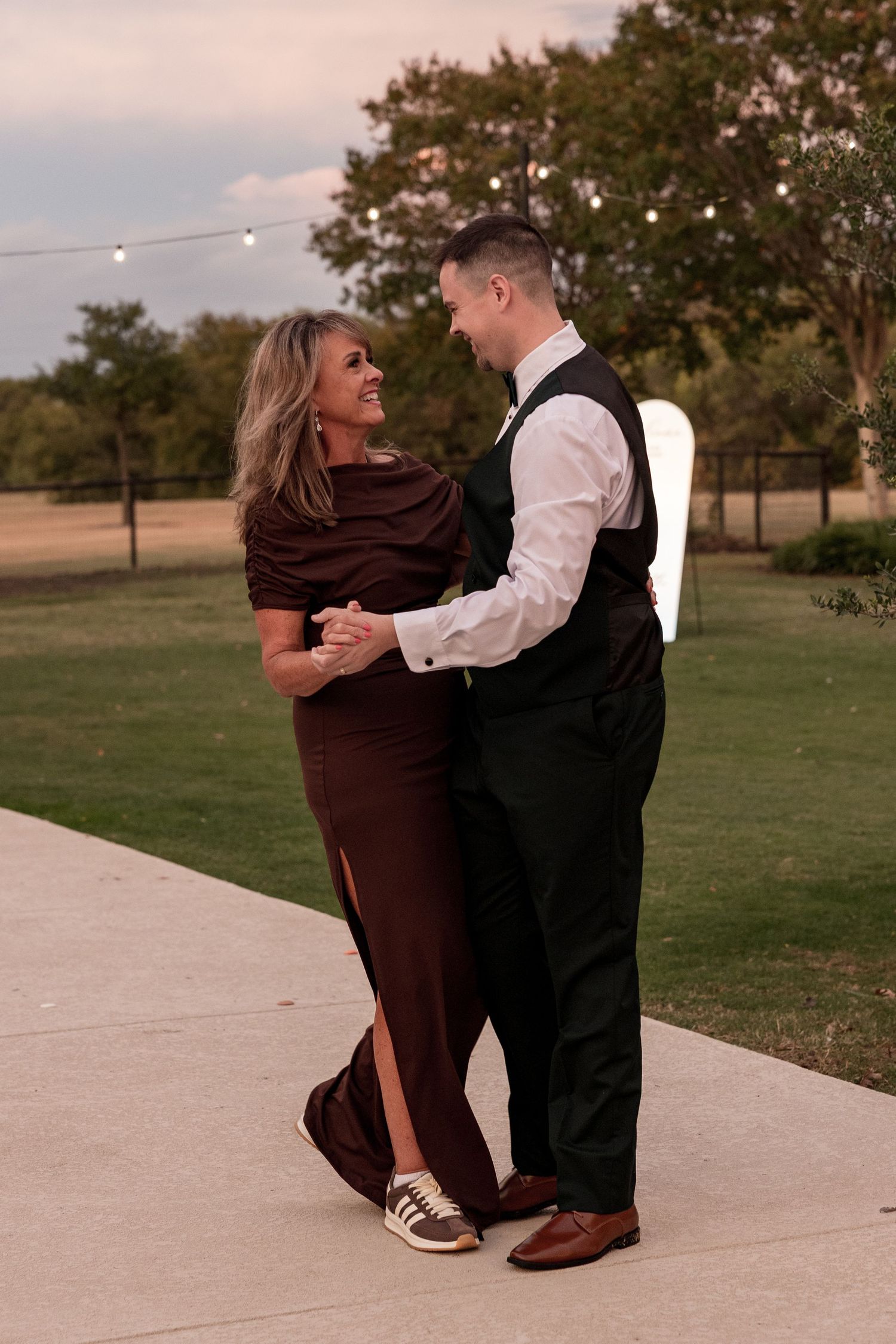 A couple in formal attire dances together on a concrete path at sunset in a grassy outdoor setting.
