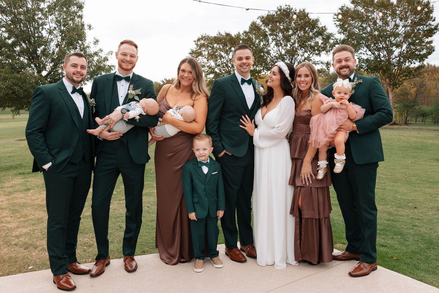 Wedding party in formal attire stands together on outdoor patio during autumn celebration.