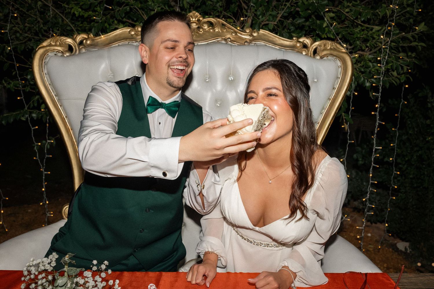 Couple shares a playful cake-feeding moment while seated on an ornate vintage sofa during their wedding celebration.