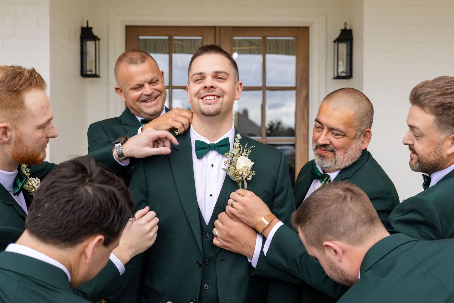 A group of groomsmen in dark green jackets share laughs and hugs during a wedding celebration.