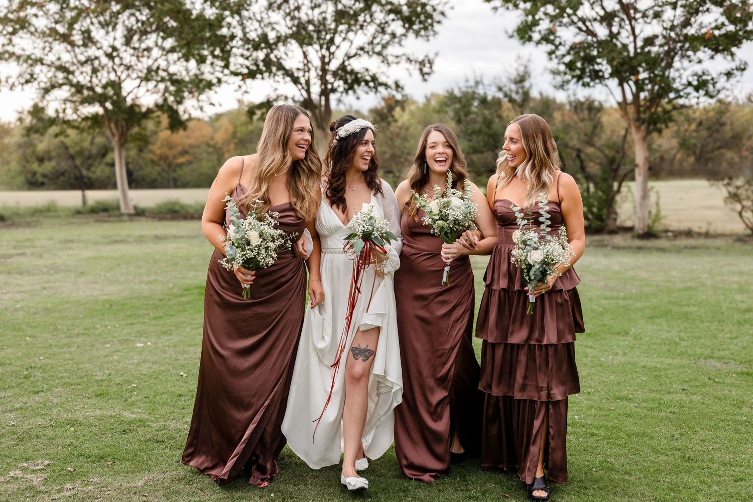 A wedding party wearing chocolate brown dresses poses together on a grassy field during an outdoor celebration.
