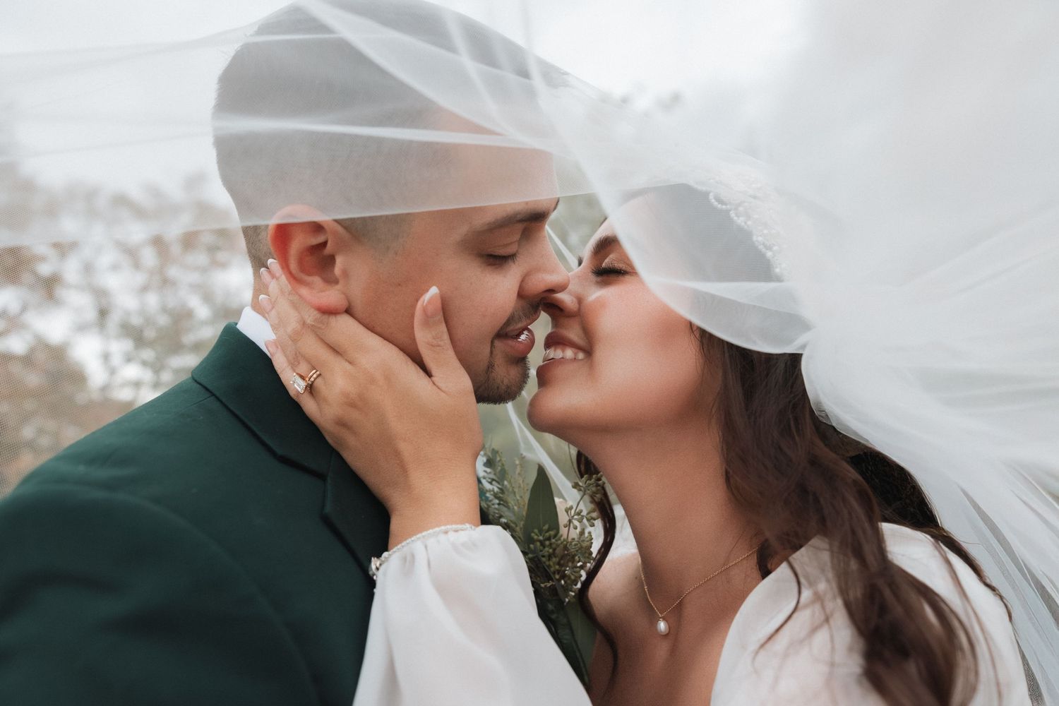 A romantic wedding couple shares a tender kiss under a flowing white veil in an outdoor setting.