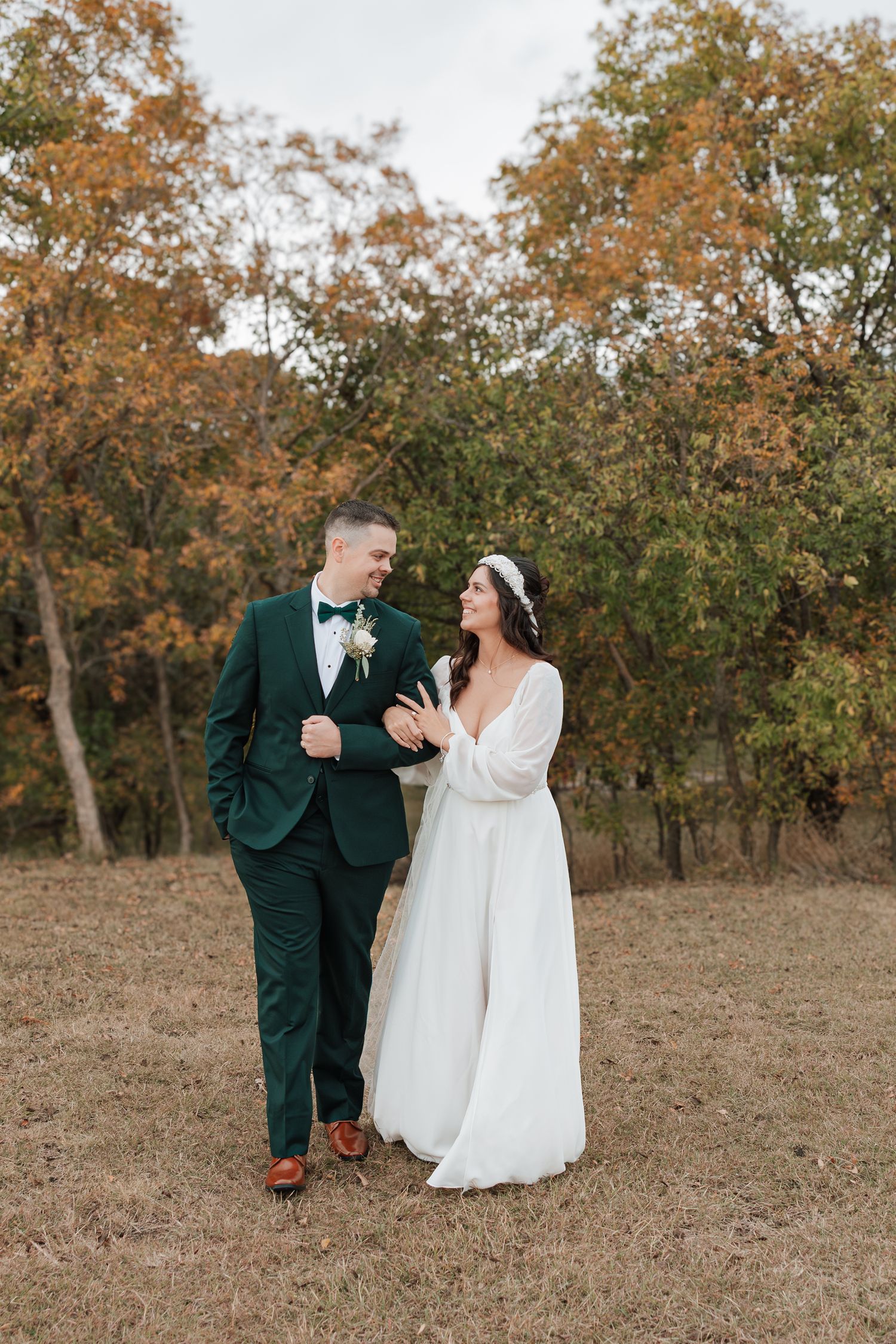 A couple in wedding attire walks together through an autumn field with golden trees in the background.