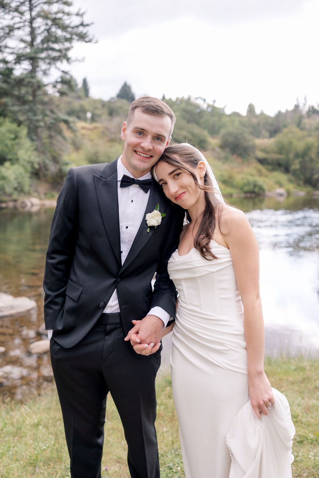 Romantic couple in formal attire poses by a scenic river with large rocks and lush greenery in the background.