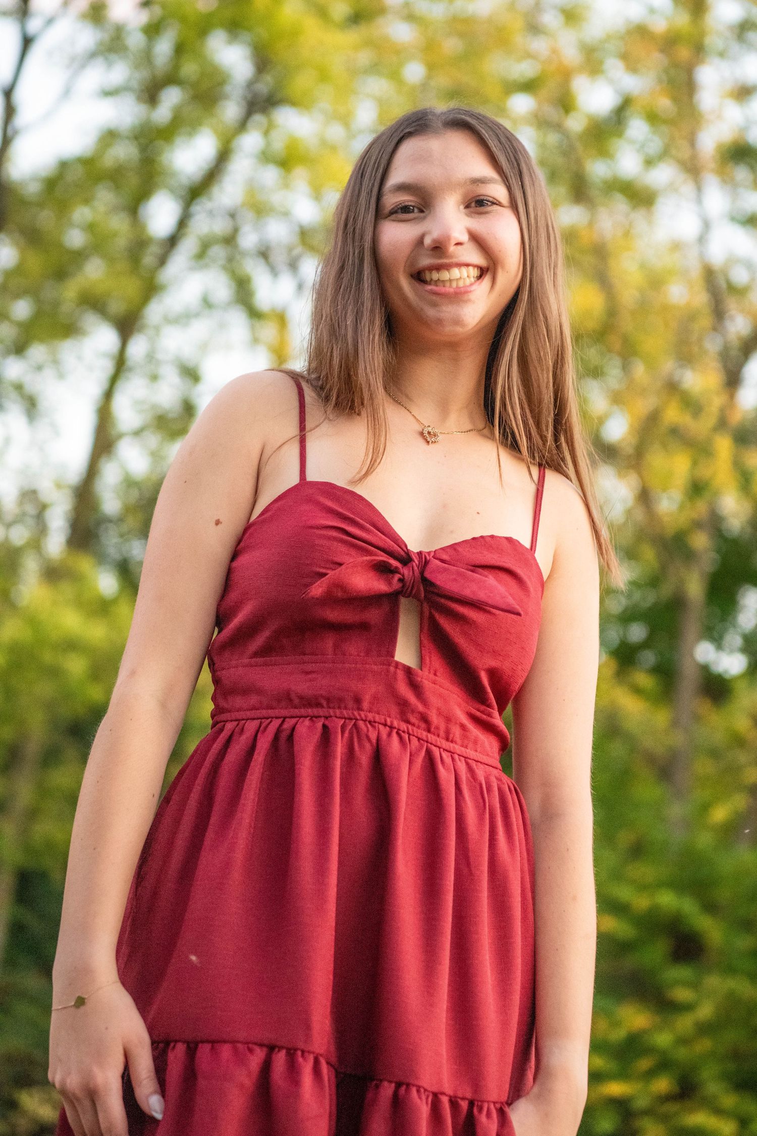 A person wearing a burgundy summer dress smiles outdoors with green foliage in the background.