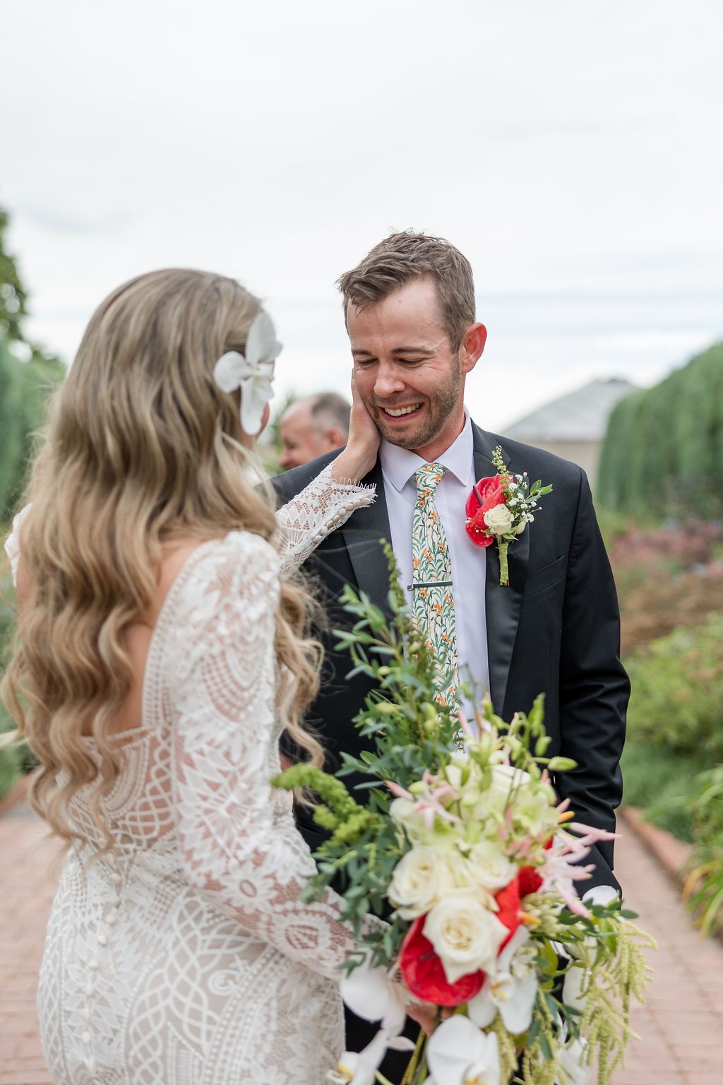 A wedding couple shares a joyful first look moment in an outdoor garden setting with red and white floral bouquet details.