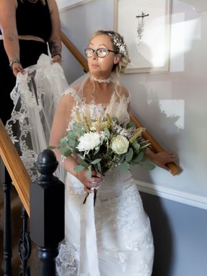 A bride in a lace wedding dress holds a white rose bouquet while standing in a brightly lit room.