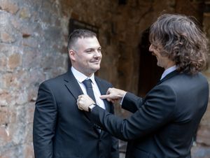 A person adjusts another person's bowtie while both wear formal black suits at a stone archway venue.