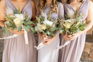 Bridesmaids in blush pink dresses holding white and green bouquets stand together at a wedding ceremony.