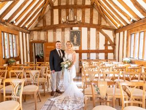 A rustic barn-style wedding venue interior with exposed wooden beams and natural lighting illuminating empty tables and chairs.