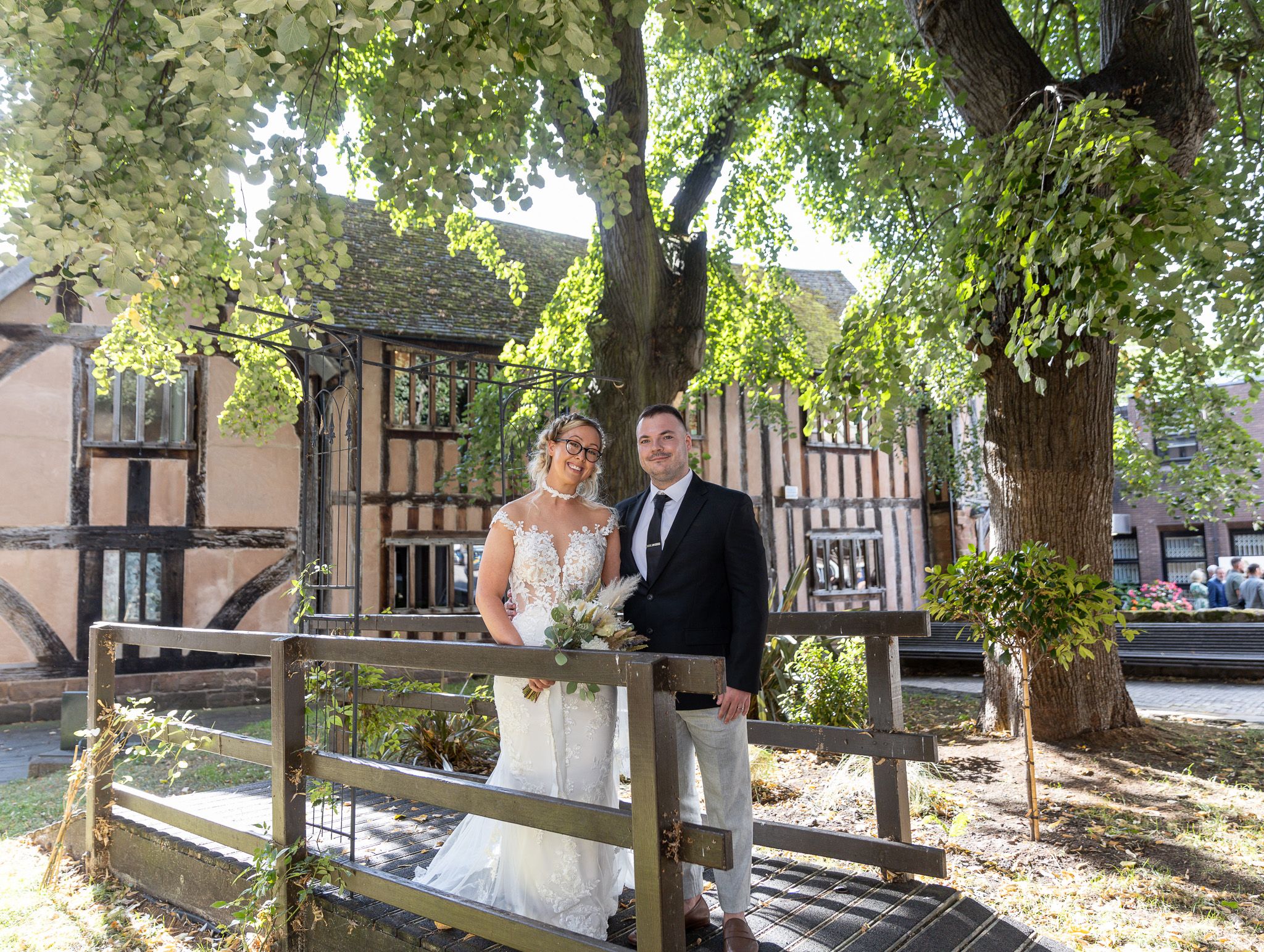 A couple walks along a tree-lined path with Tudor-style buildings visible in the background on their wedding day.