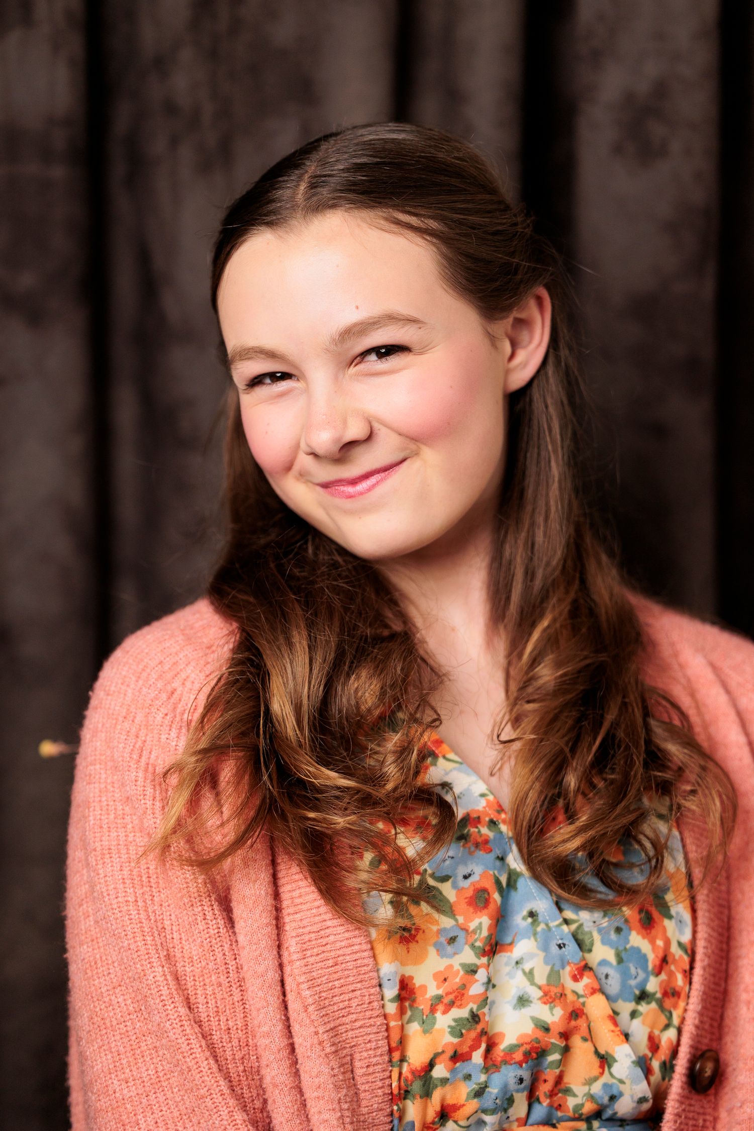 A teenage girl with long brown hair smiles warmly wearing a pink cardigan over a floral dress.