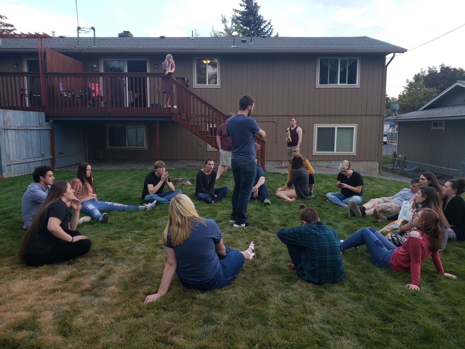 Large group of college students sitting in a circle on a backyard lawn during an outdoor group activity.