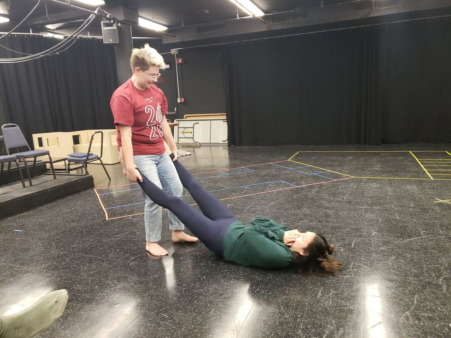 Two performers rehearsing an improv comedy scene on a black studio floor with one person lying down.
