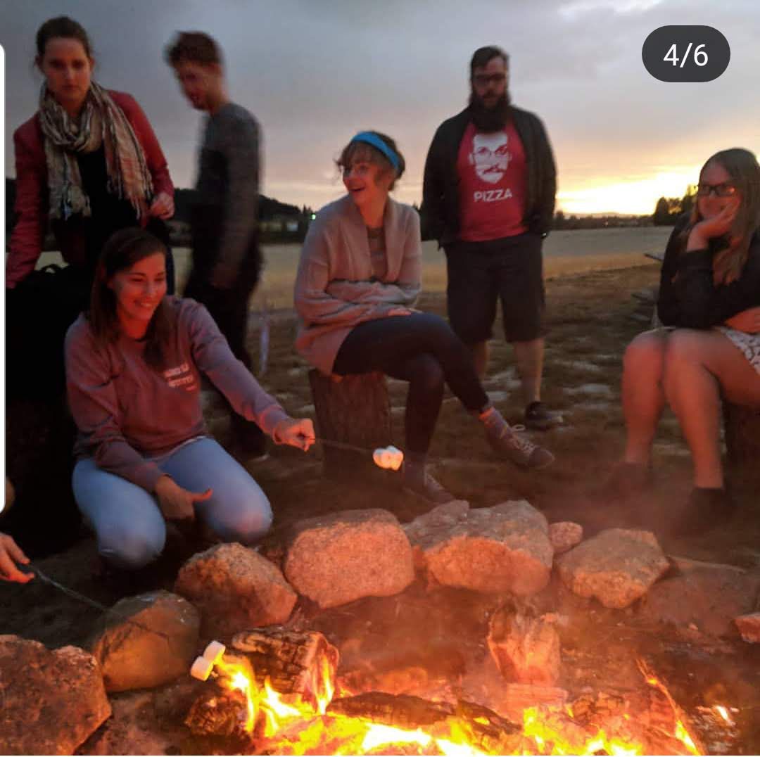 Group of young adults roasting marshmallows around a stone fire pit at sunset on a beach.