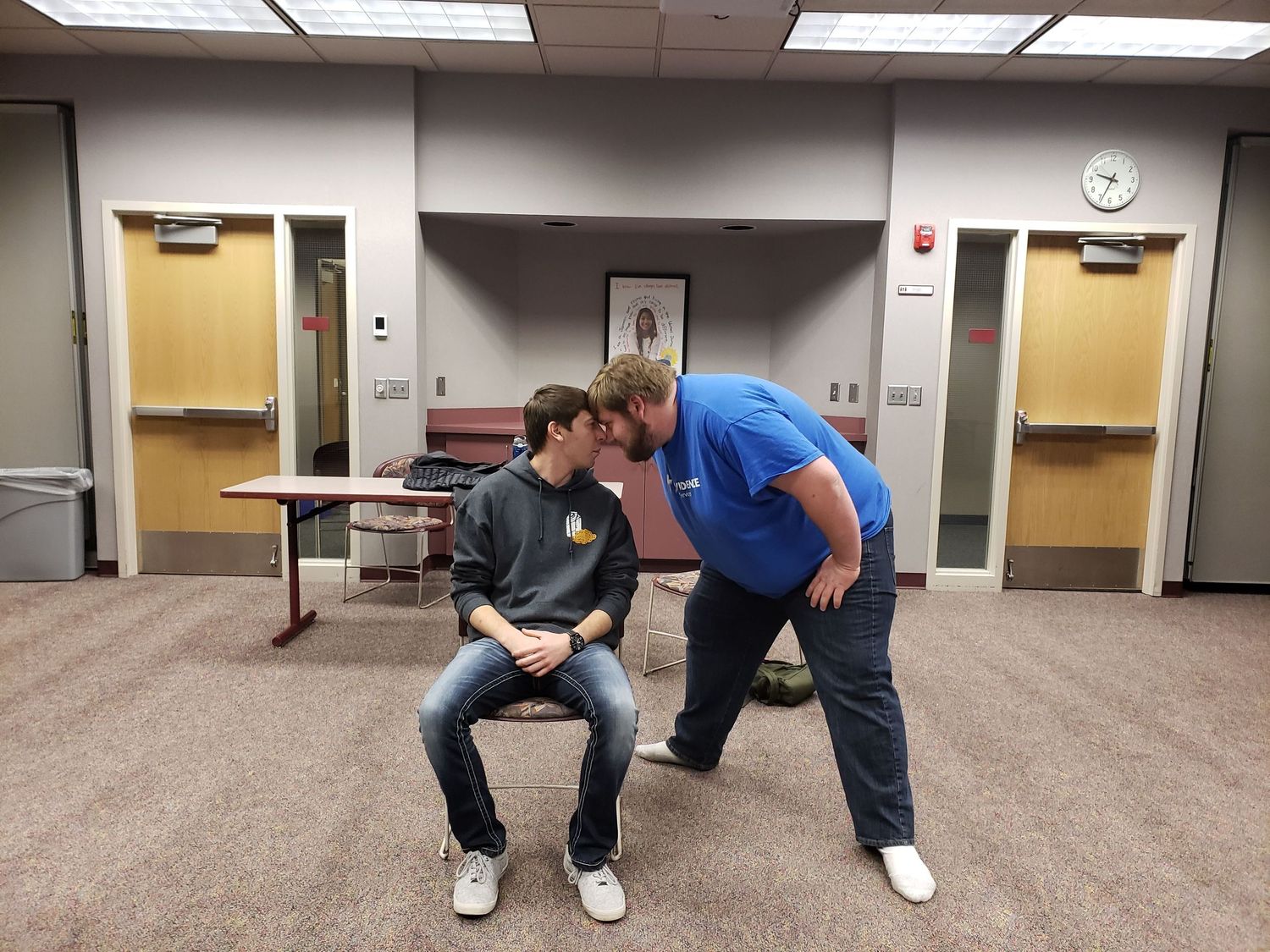 Two young men rehearsing an improv comedy scene in a classroom, leaning close face to face.