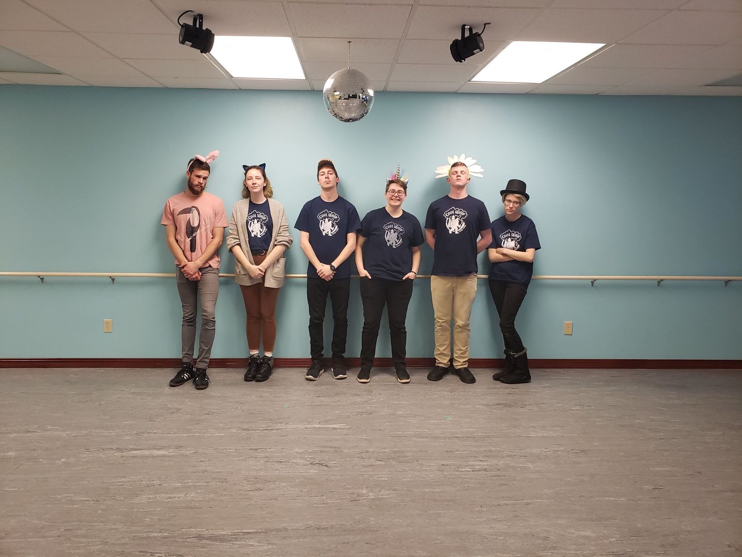 Six performers in matching navy blue t-shirts posing in a dance studio with blue walls and disco ball.
