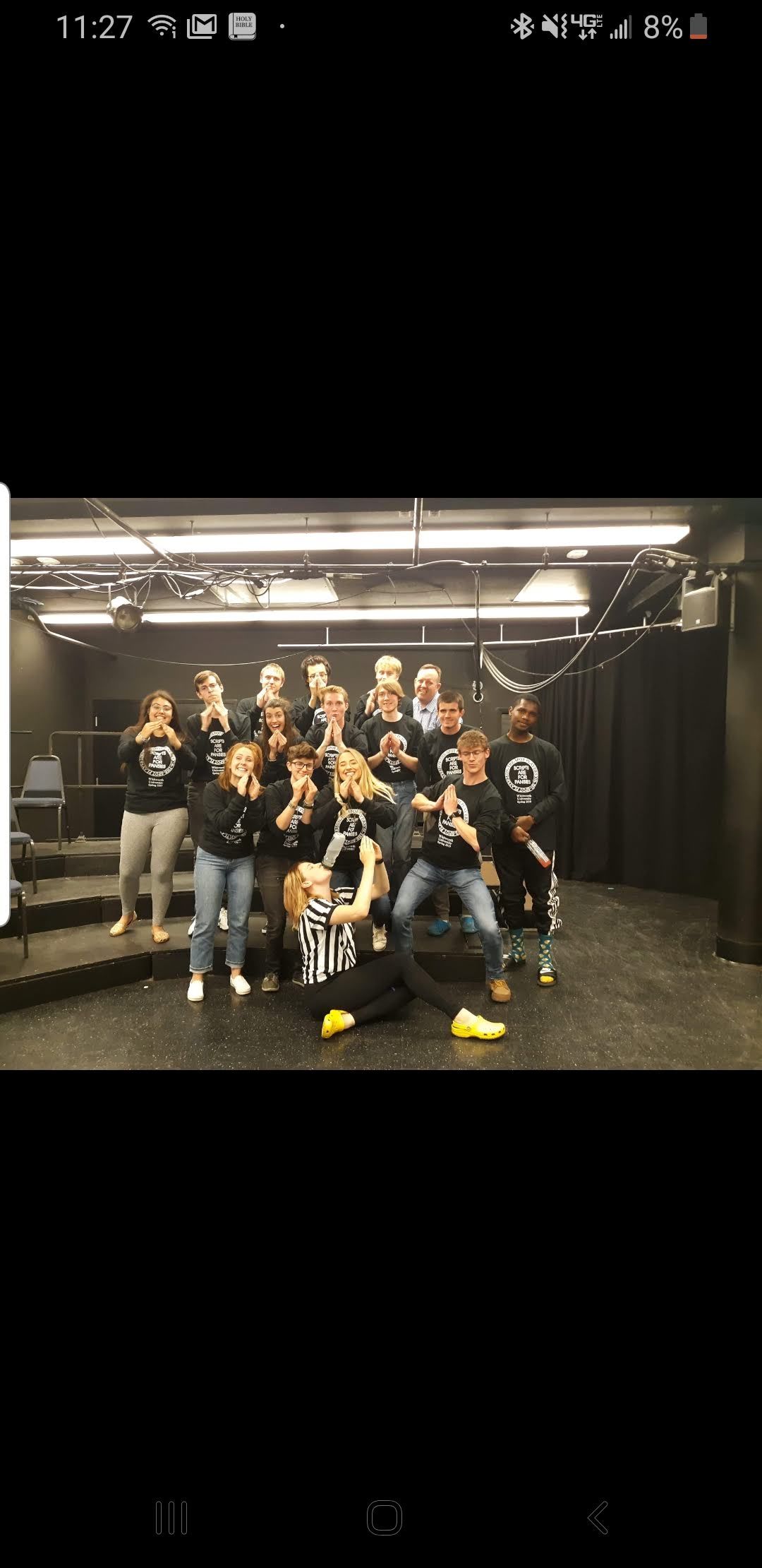 Group of performers in matching black t-shirts posing together backstage in a black box theater.