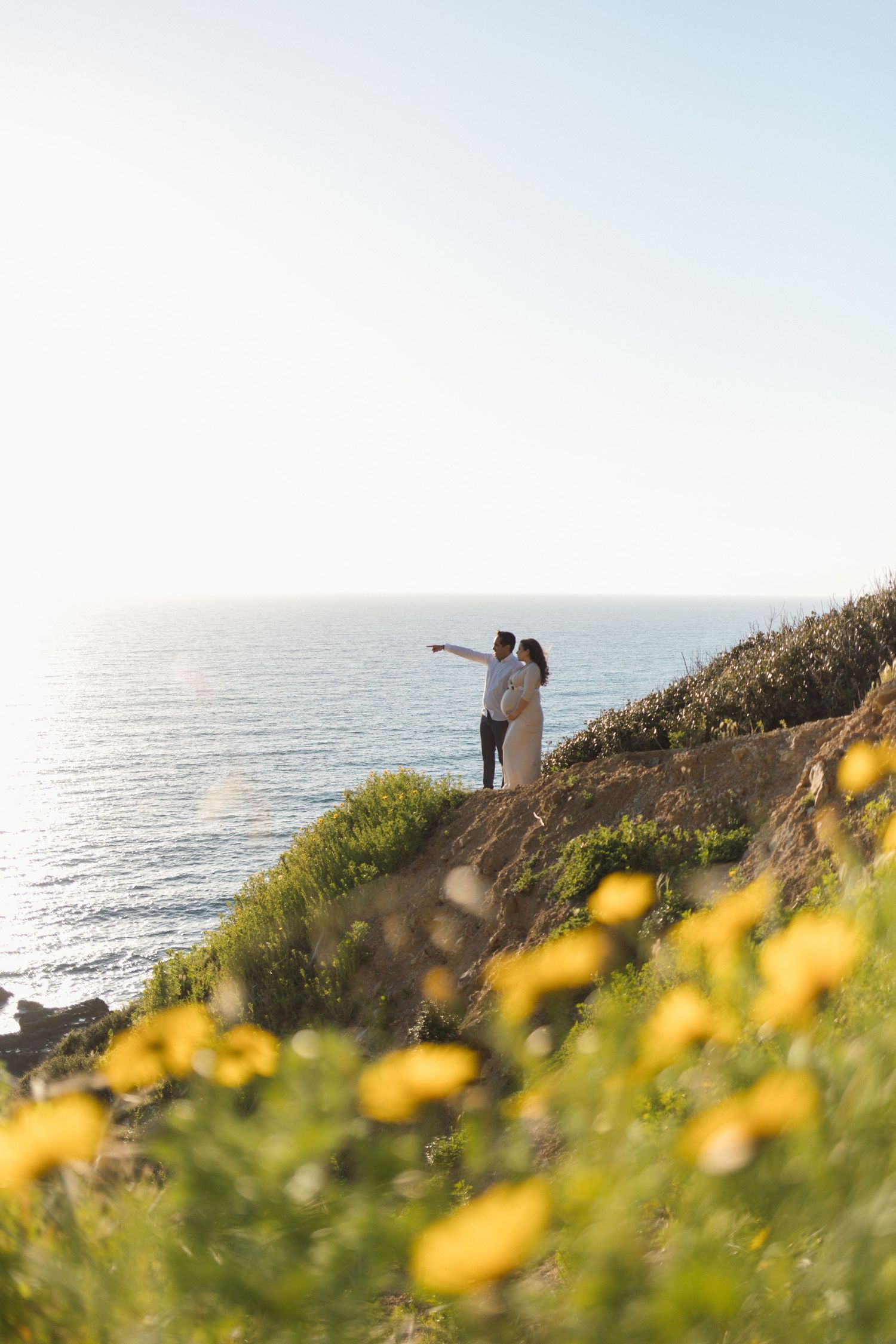 Maternity session on the Southern California coast with a couple overlooking the ocean and yellow flowers in the foreground