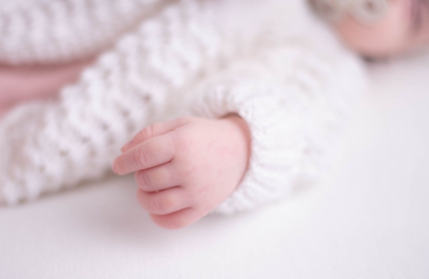 Tiny newborn baby hand in white knit romper sleeve, close-up infant photography.