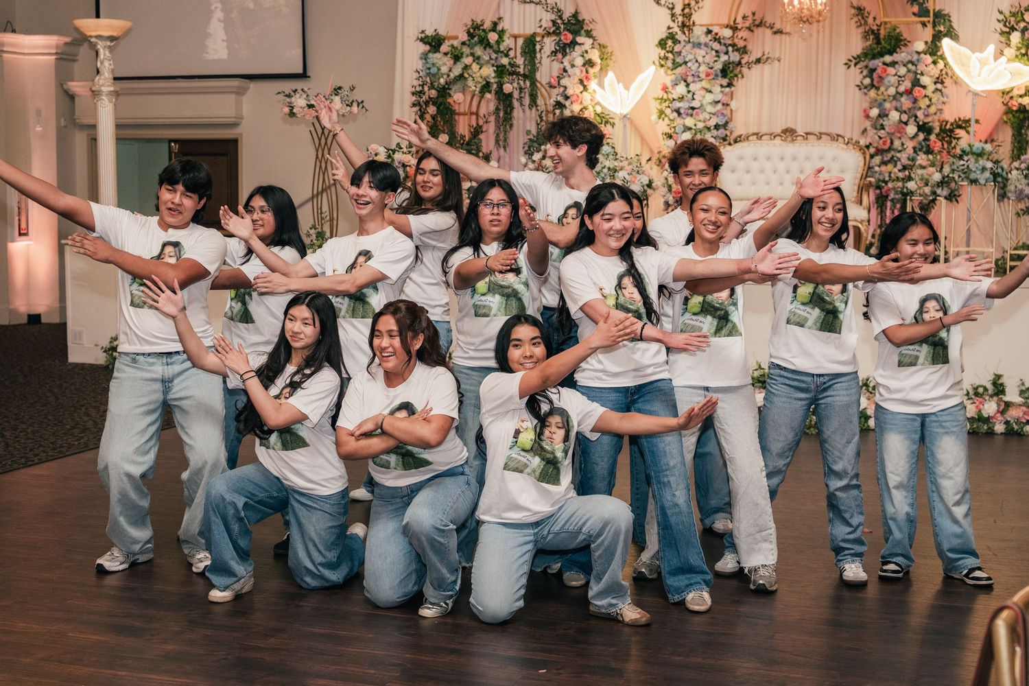 Group of teens in matching white graphic tees and jeans posing playfully at a floral decorated event venue.