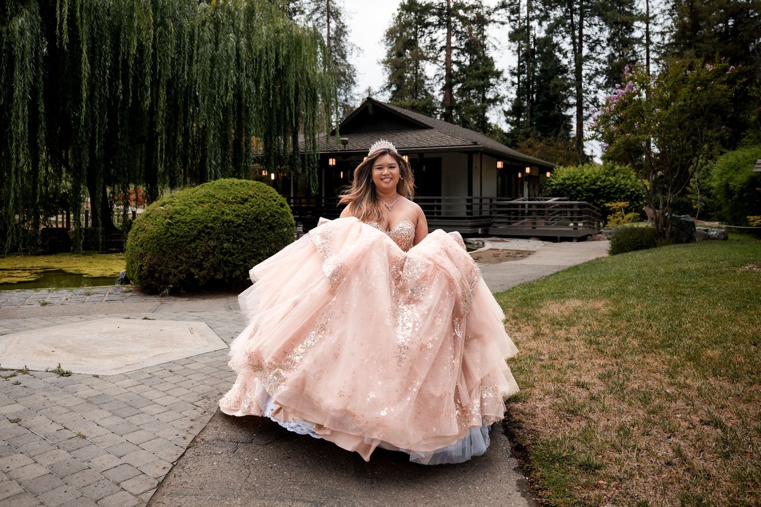 Young woman twirling in a blush pink ball gown with floral details in a lush garden setting.