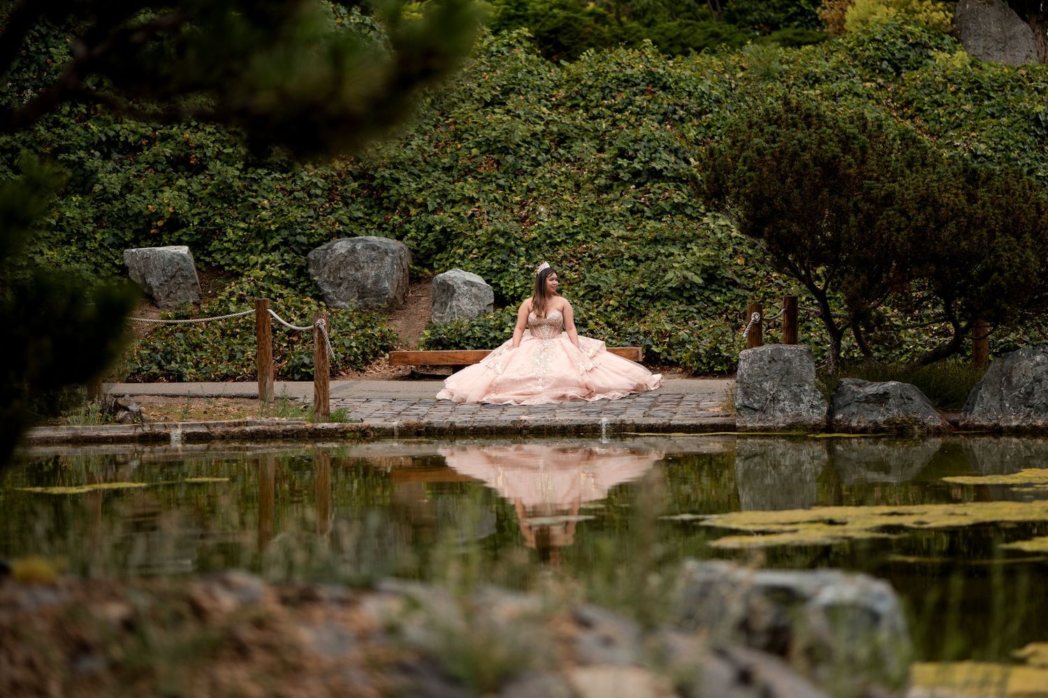A woman in a flowing pink gown sits gracefully by a serene Japanese garden pond surrounded by lush greenery.