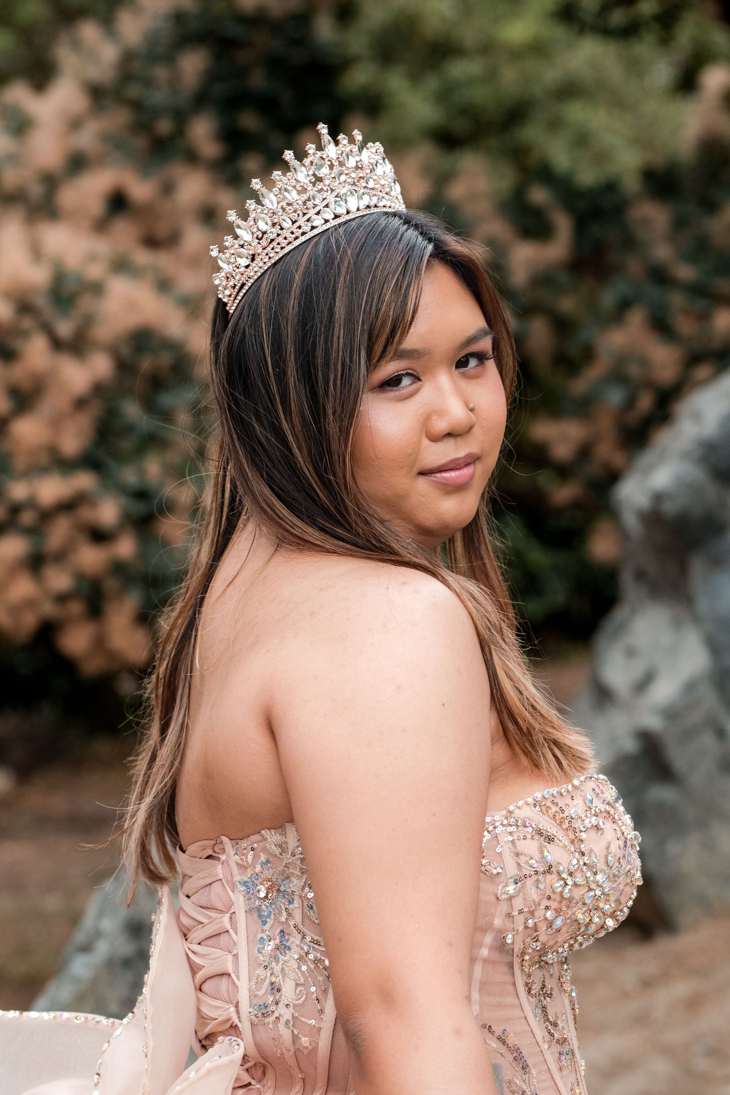 Young woman in a beaded blush gown and crystal tiara poses outdoors for a quinceañera portrait.