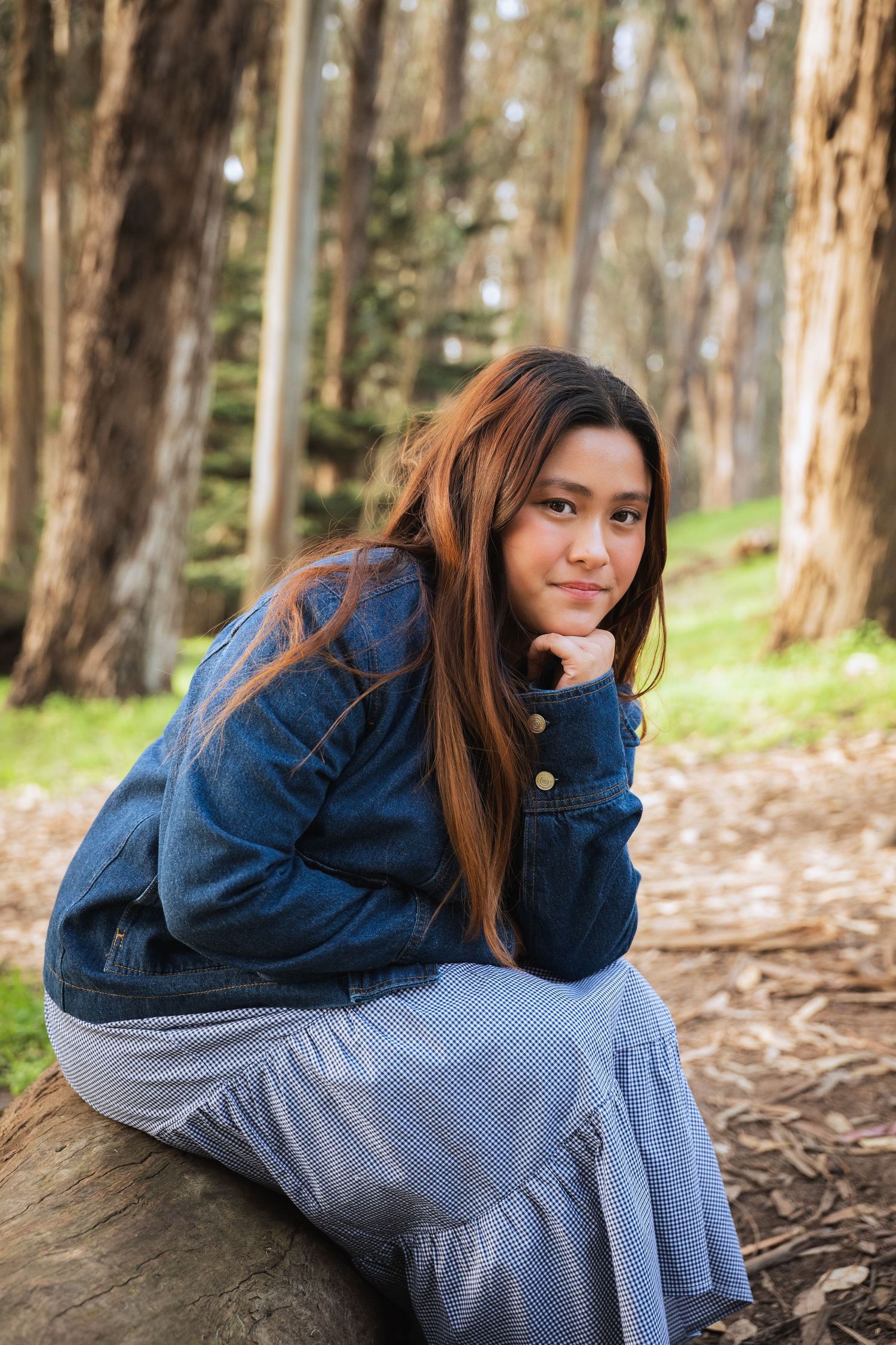 Young woman in denim jacket and blue skirt sits on a log in a forest, smiling with chin resting on hand.