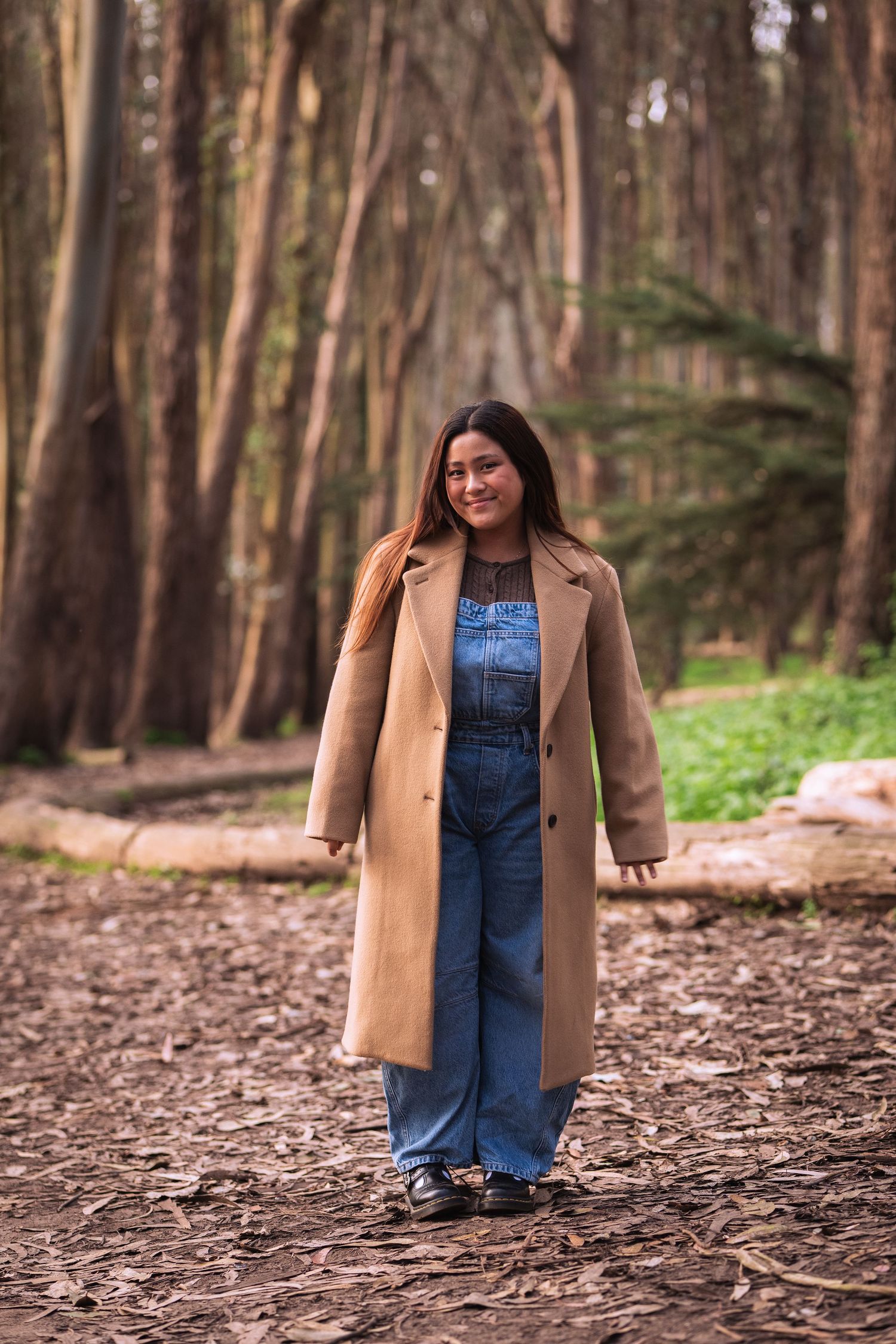 Person in denim overalls and tan coat poses in a wooded forest setting with fallen leaves and pine trees.